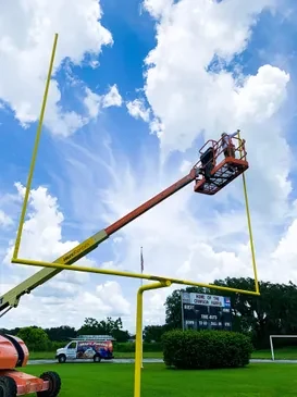 Painter on a boom lift repainting a yellow goalpost on an athletic field