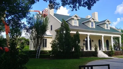 Painter working from a boom lift on the upper storey of a large cream-colored home with a green metal roof